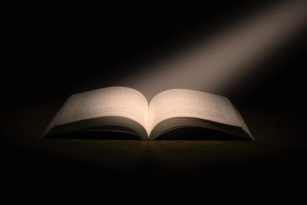 Un rayo de luz incide sobre un libro abierto, aproximadamente por el centro de sus páginas. El entorno es oscuro para resaltar la iluminación sobre el libro.
