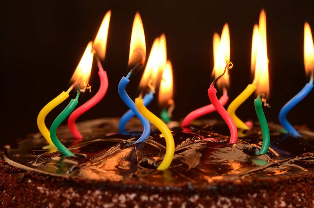 Zoom sobre una tarta de cumpleaños, sobre un fondo negro. Solo se ve la capa superior de esta y las velas encendidas. Estas son de colores y aparecen deformadas por el calor de la llama. La tarta parece de chocolate.